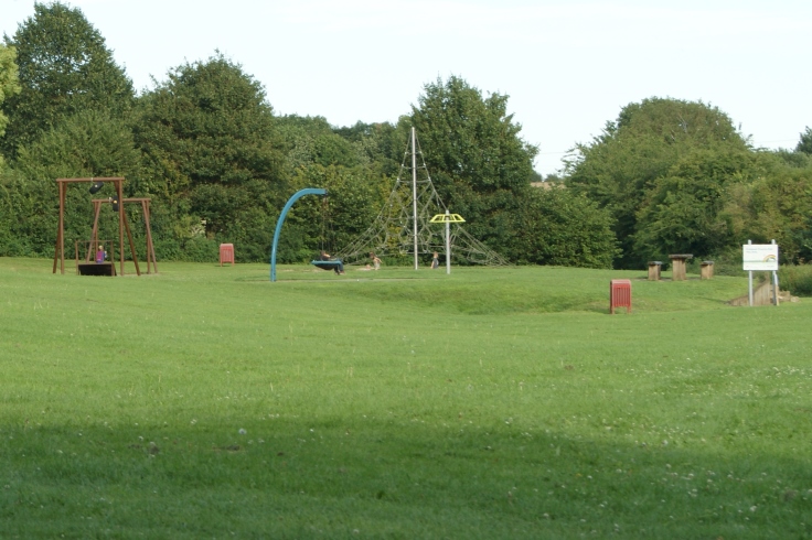 A park with a play area, bench, picnic area, bin and a sign for the play area.